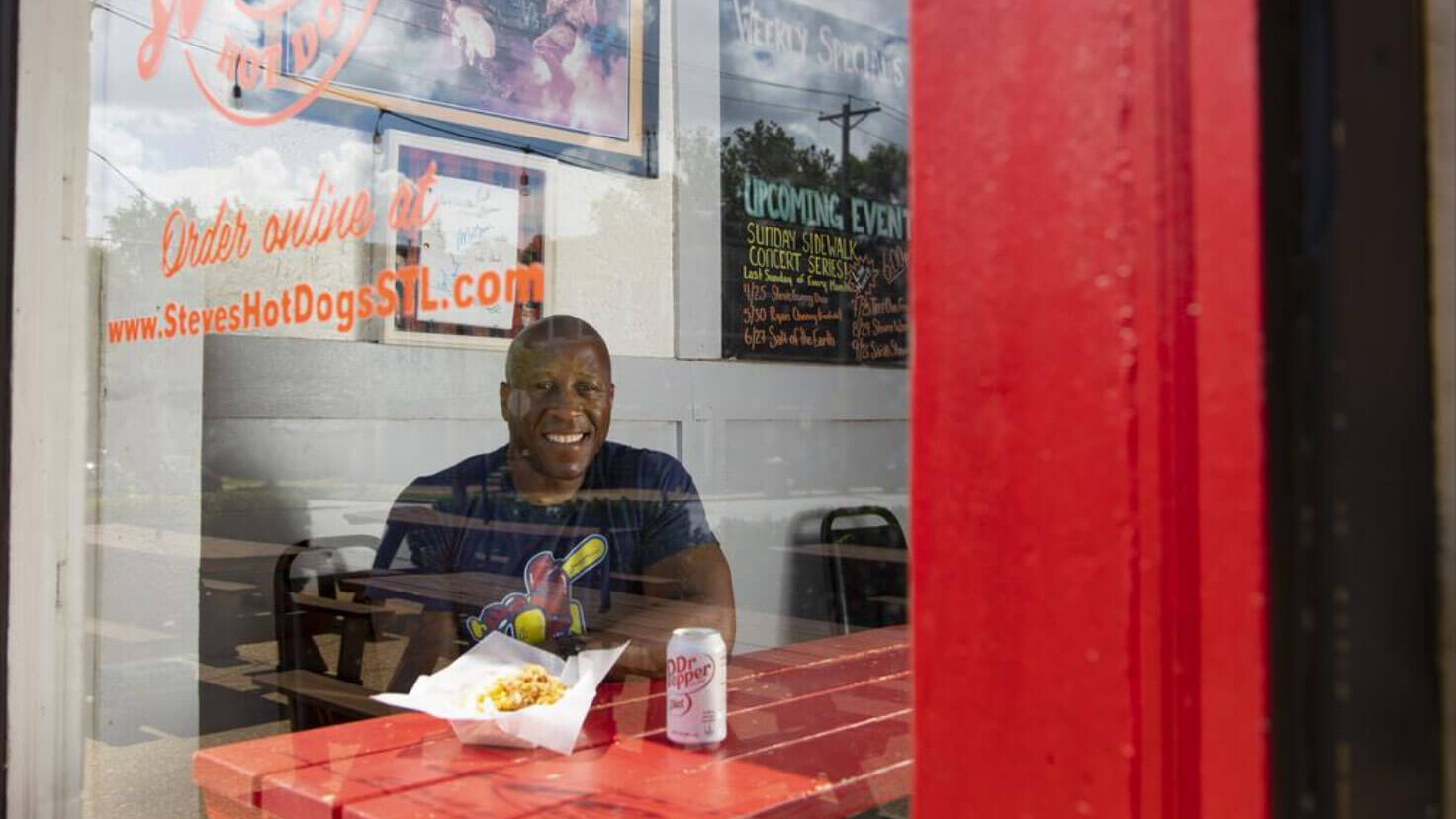 A smiling man sitting behind a glass window and facing the camera.