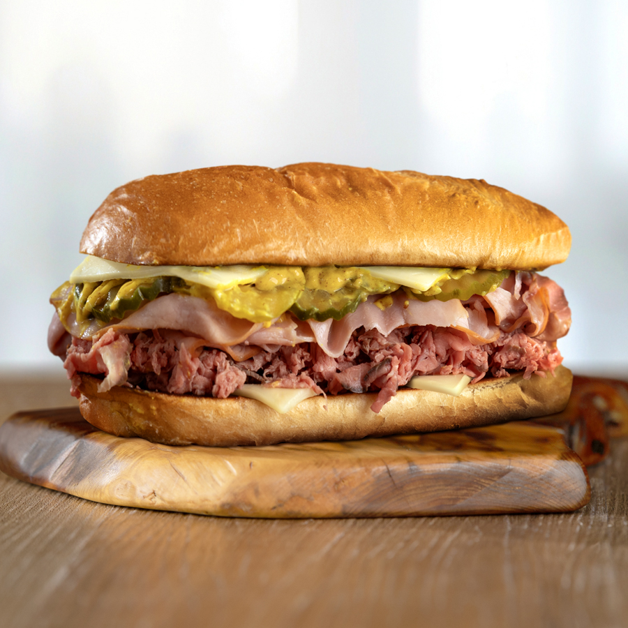 A sandwich on a wood surface. On a hoagie bun with visible meats, cheese, pickles, mustard