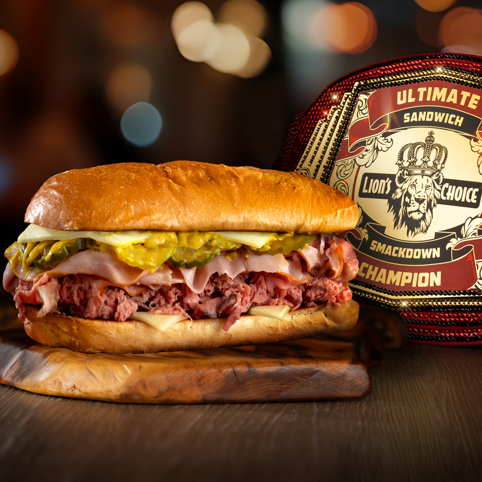 A photograph of a large meat and cheese and pickle sandwich on a hoagie bun. The sandwich is sitting in front of a championship belt