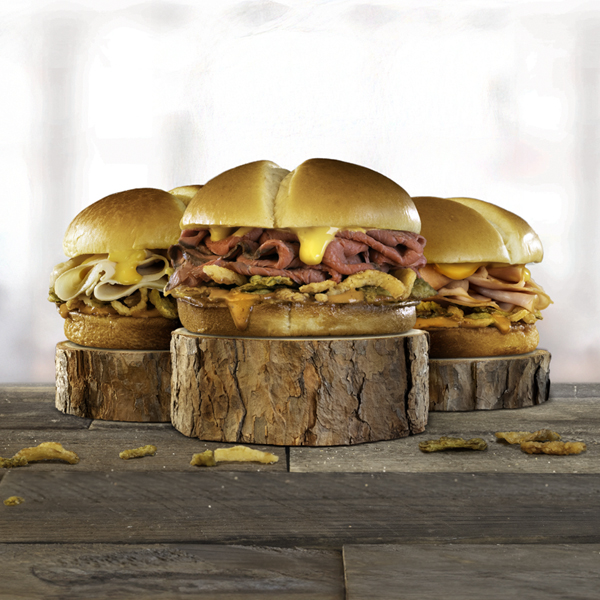 Image of three sandwiches on buns with different meat and cheeses. They are each sitting on a circular slice of a wood log